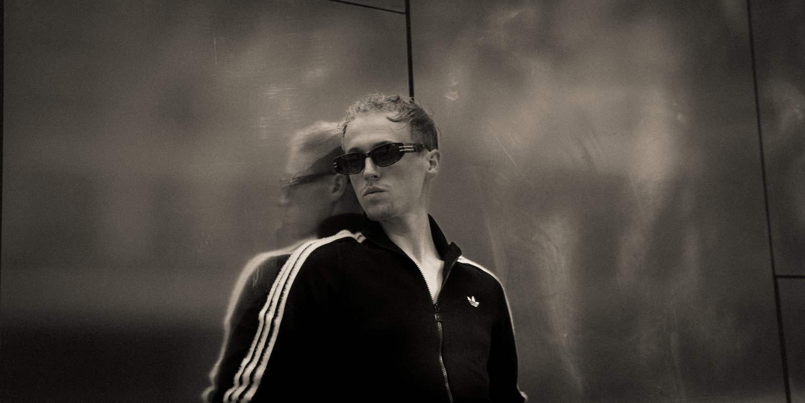 Léo Martin en noir et blanc, lunettes de soleil, son reflet dans un mur de métal